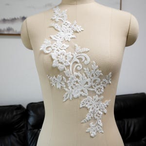 May include: A white lace applique featuring an intricate floral pattern with embroidered leaves and small pearl accents. The design is arranged in an elongated, asymmetrical shape, crafted from delicate white thread on a sheer mesh base.