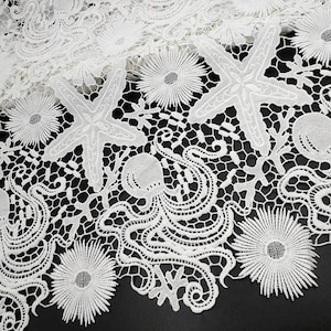 Op de afbeelding: Close-up van witte kanten stof met een zeelevenontwerp. Het kant heeft zeesterren, koraal en andere mariene elementen. De ingewikkelde details en het open werk creëren een delicate en elegante textuur.