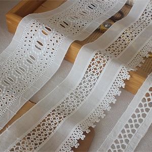 May include: Three white cotton lace trims with different patterns. The lace trims are arranged on a wooden surface.