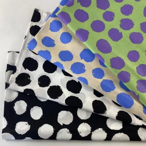 May include: Three folded pieces of fabric featuring a large, hand-painted style polka dot pattern. The top layer is light green with purple dots, the middle is beige with blue dots, and the bottom layer is white with large black circular spots.