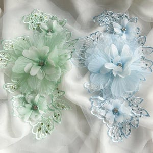 May include: Two floral lace appliques, one in mint green and one in light blue, featuring layered fabric petals, intricate embroidered leaf patterns, and small clear rhinestones centered on each flower and scattered along the lace edges.