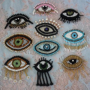 May include: A collection of beaded eye-shaped brooches in various colors and designs. The brooches feature intricate beadwork, including pearls and colorful beads, with some having dangling embellishments. The eyes are in shades of black, white, gold, green, and blue.