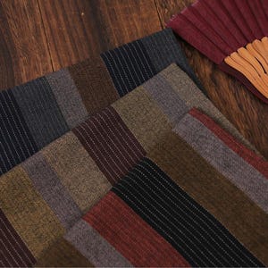以下が含まれることがあります： グレー、ブラウン、バーガンディなど、さまざまな色の縦縞模様の生地見本をクローズアップ。各見本には、細い白いアクセントストライプがあります。背景には、部分的に見えるバーガンディの扇子があります。