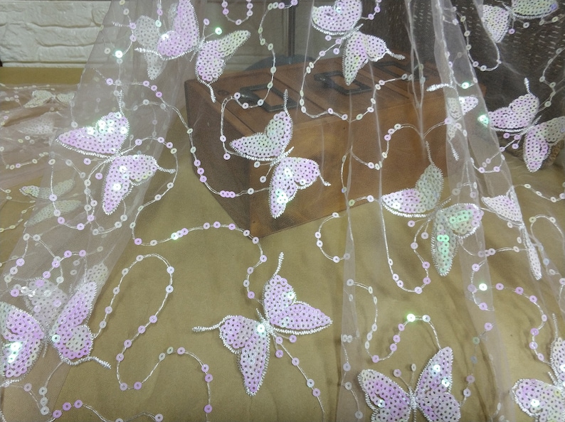 May include: White sheer fabric with pink butterfly sequins. The fabric is delicate and has a soft, flowing texture.