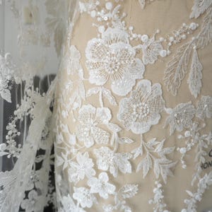 May include: A sheer white fabric featuring intricate floral embroidery with white thread, small clear sequins, and tiny white beads. The floral pattern covers the mesh material, creating a textured, layered appearance on a beige dress form.