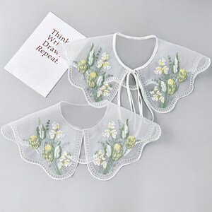 Op de afbeelding: Twee witte, doorschijnende, geborduurde kragen met geschulpte randen en bloemmotieven. De kragen hebben gele en witte bloemen met groene bladeren. Eén kraag heeft een striksluiting. Een wit papier met de tekst "Think, Write, Draw, Repeat" staat op de achtergrond.