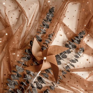 May include: A brown paper pinwheel with a white center sits on a brown tulle fabric with gold glitter stars. Dried eucalyptus branches are scattered around the pinwheel.