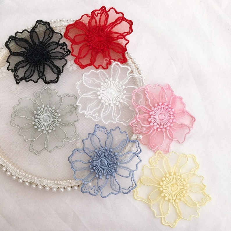 May include: A collection of seven sheer floral appliques in black, red, white, pink, light blue, yellow, and grey. Each flower features intricate embroidered edges and a detailed center, arranged on a circular white lace base with pearl trim.