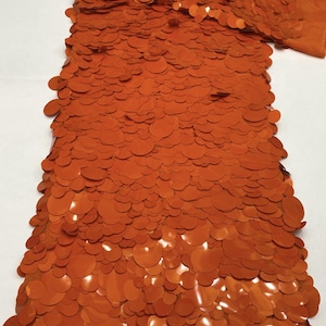 Puede incluir: Una muestra de tela de color naranja vibrante, densamente cubierta con lentejuelas circulares superpuestas de varios tama&ntilde;os. Las lentejuelas est&aacute;n unidas a una tela base naranja a juego, creando una superficie texturizada y reflectante con la palabra Orange.