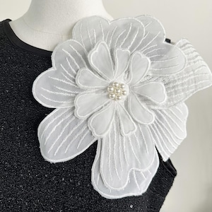 以下が含まれることがあります： 繊細な刺繍が施された、中央に模造真珠のクラスターが付いた、大きな白い花のアップリケ。 アップリケは重ねられており、透け感のある生地で作られているようです。 花は黒い衣服に取り付けられています。