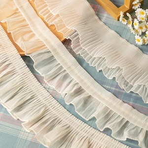 Peut inclure: Gros plan de plusieurs garnitures de tissu à volants délicats dans des tons de blanc et de crème. Les garnitures varient en largeur et en style de volants, avec un aspect doux et fluide. Le tissu semble léger, peut-être de la mousseline ou de l'organza, adapté aux projets de couture et d'artisanat.