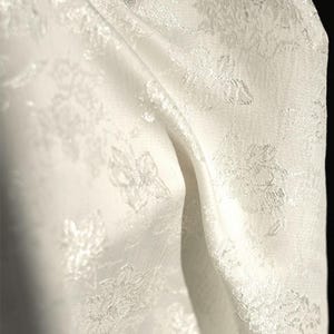 Könnte beinhalten: Nahaufnahme eines weißen Stoffes mit floralem Muster. Der Stoff ist drapiert und zeigt die Textur und das Design der gestickten Blumen. Das Material wirkt weich und zart, mit dezentem Glanz.