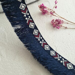 May include: A dark blue woven trim featuring a long fringe along one edge and a repeating geometric pattern in white and red thread. The design consists of diamond shapes and chevron accents embroidered along the length of the fabric ribbon.