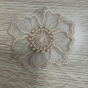 May include: A beige floral applique patch featuring intricate embroidery on a sheer, translucent mesh fabric. The design showcases a central circular cluster of raised stitching surrounded by five distinct, scalloped petals with detailed thread borders.