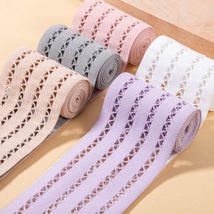 Peut inclure: Cinq rouleaux de ruban élastique dans des tons pastel rose, gris, pêche, lavande et blanc. Chaque rouleau présente un motif ajouré répétitif avec des bandes tissées verticales et une série de petites découpes triangulaires sur toute la longueur.