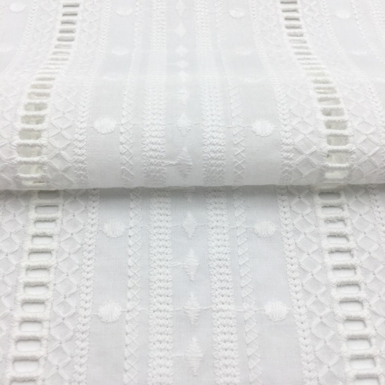 May include: White cotton fabric with embroidered vertical stripes of different patterns. The fabric has a delicate, lacy look.