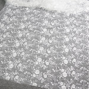 Puede incluir: Tela de encaje floral blanco con un delicado patr&oacute;n de flores y hojas. El encaje est&aacute; detallado con adornos florales en relieve. La tela est&aacute; sobre una superficie gris y es semitransparente.