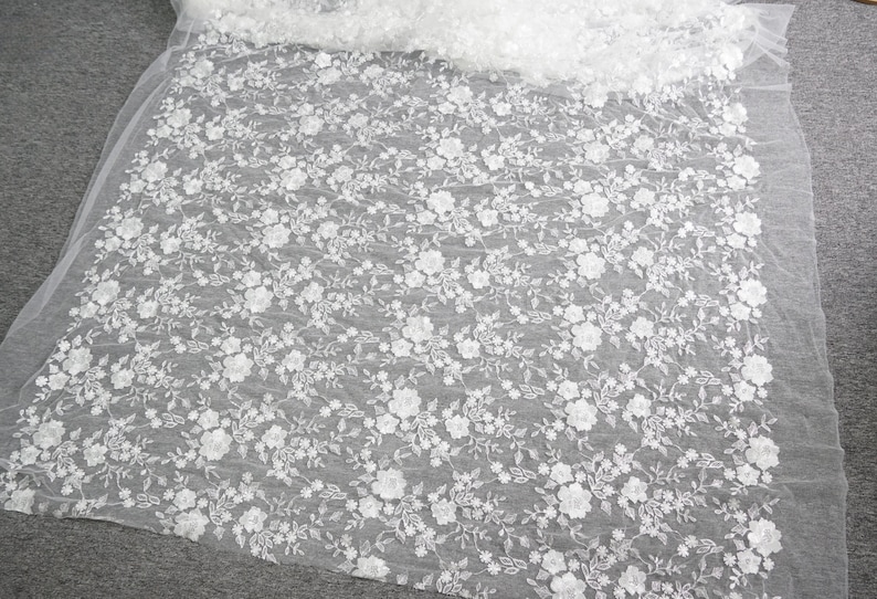 Puede incluir: Tela de encaje floral blanco con un delicado patr&oacute;n de flores y hojas. El encaje est&aacute; detallado con adornos florales en relieve. La tela est&aacute; sobre una superficie gris y es semitransparente.