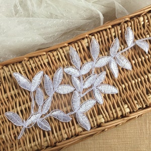 May include: White lace applique with a delicate floral design featuring leaves and branches. The applique is displayed on a woven wicker surface.