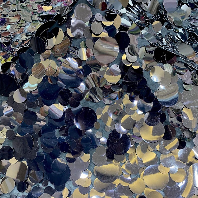 Puede incluir: Una superficie de tela densa cubierta con lentejuelas met&aacute;licas circulares superpuestas en tonos plateado, gris plomo y gris oscuro. Las lentejuelas var&iacute;an en tama&ntilde;o y est&aacute;n dispuestas en capas, creando una apariencia altamente reflectante y texturizada.