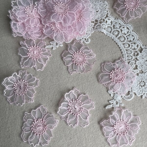 May include: A collection of light pink floral lace appliques featuring sheer petals with embroidered edges and centers. The set includes individual flower motifs and a larger cluster, accompanied by white lace trim with a scalloped edge pattern.