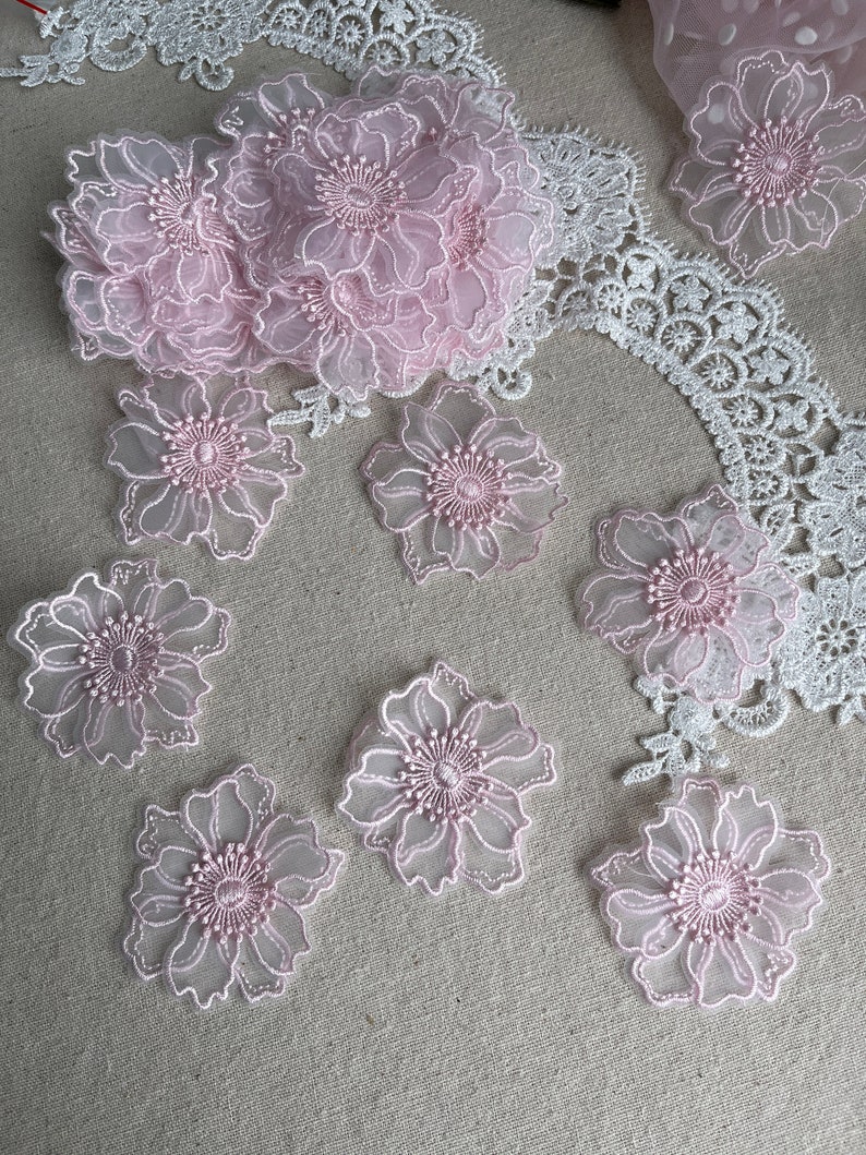 May include: A collection of light pink floral lace appliques featuring sheer petals with embroidered edges and centers. The set includes individual flower motifs and a larger cluster, accompanied by white lace trim with a scalloped edge pattern.