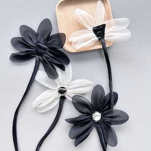 May include: Four fabric flower hair accessories in black and white. The flowers have a daisy-like design with petals and long, slender stems. Some flowers have a decorative gem in the centre. The flowers are arranged on a light grey surface.