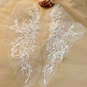Könnte beinhalten: Zwei weiße florale Spitzenapplikationen mit Perlen- und Paillettenbesatz. Die Applikationen sind auf einem durchsichtigen Netzstoff.