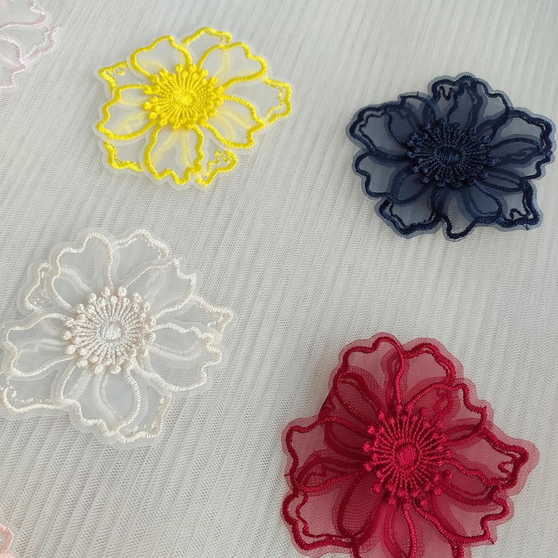May include: Four floral appliques made of sheer organza fabric with intricate embroidery. The set includes one bright yellow flower, one dark navy blue flower, one off-white flower, and one deep red flower, all featuring detailed centers.