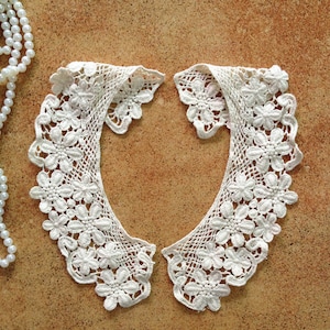 May include: A pair of white crocheted lace collars with a floral design. The collars are laid out on a brown surface.