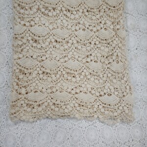 May include: A close-up of a beige crocheted lace fabric with a wavy pattern. The fabric is folded in half and is lying on a white crocheted lace fabric.