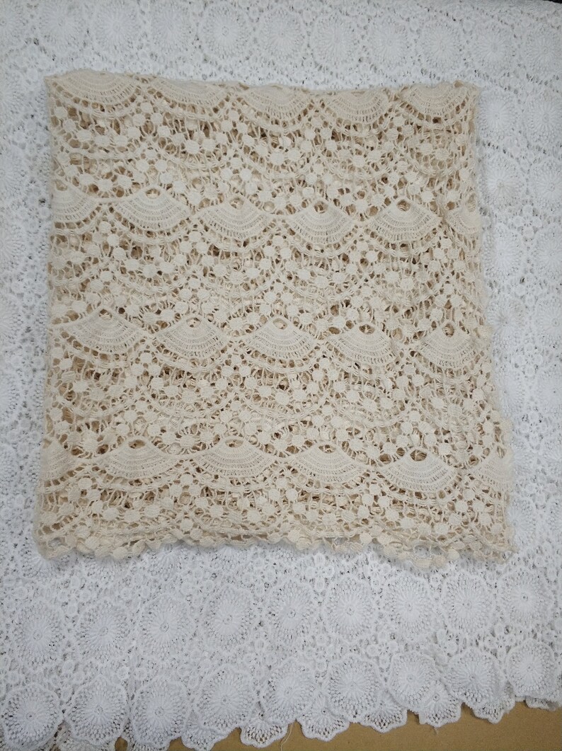 May include: A close-up of a beige crocheted lace fabric with a wavy pattern. The fabric is folded in half and is lying on a white crocheted lace fabric.
