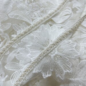 May include: White lace fabric with a beaded trim. The trim is made of small, white beads that are sewn together in a continuous line.