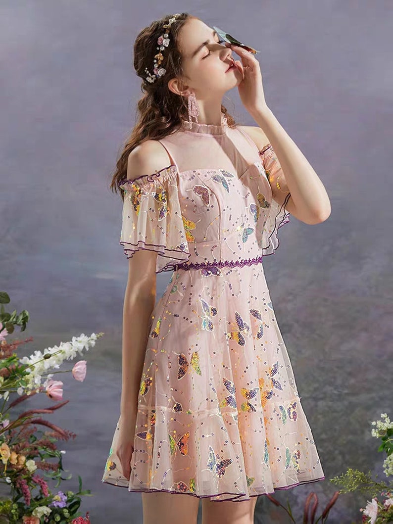 May include: A pink off-the-shoulder dress with butterfly sequins. The dress has a ruffled neckline and short sleeves.
