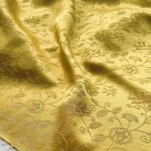 May include: Close-up of a golden yellow fabric with a floral pattern. The fabric has a subtle sheen and is draped, creating shadows that highlight the embossed floral design. The pattern includes flowers, leaves, and winding stems.