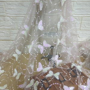 May include: Pink sheer fabric with iridescent butterfly sequins. The fabric is on a white mannequin form and has a light pink background.