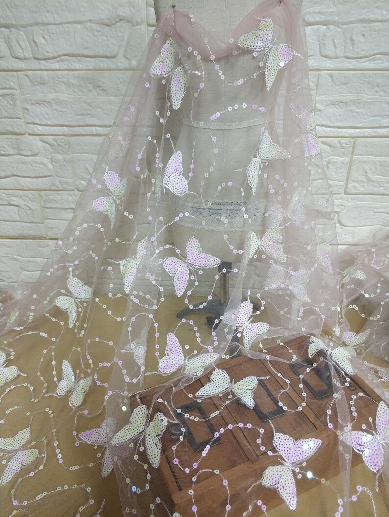 May include: Pink sheer fabric with iridescent butterfly sequins. The fabric is on a white mannequin form and has a light pink background.