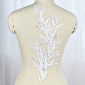 May include: A white embroidered applique featuring a detailed botanical design of elongated leaves and stems. The intricate needlework creates a textured, leafy pattern that is centered on a beige dress form, showcasing the embroidery's shape.