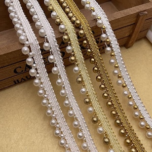 Puede incluir: Primer plano de un ribete decorativo en varios colores. El ribete presenta una base tejida con perlas de imitación colgantes y cuentas doradas. Los colores incluyen blanco, dorado y plateado, adecuados para adornar ropa o manualidades.
