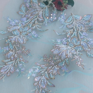 Może przedstawiać: Zbliżenie delikatnych aplikacji z koronki kwiatowej w odcieniach niebieskiego, lawendy i srebra. Haft zawiera skomplikowane koraliki i cekiny, tworząc efekt połysku. Koronka jest osadzona na jasnoniebieskim tle z tiulu, idealna do sukni ślubnych lub formalnych.