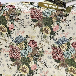 Könnte beinhalten: Cremefarbene Blumentapete mit einem sich wiederholenden Muster aus Rosen, Hortensien und anderen Blumen in Rosa, Blau, Gelb und Burgunder. Der Stoff hat eine gewebte Textur und eignet sich für die Inneneinrichtung.