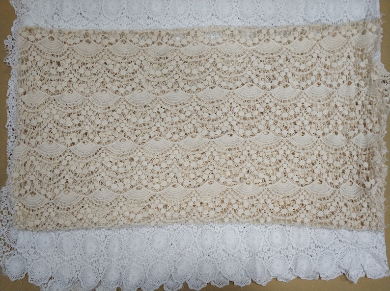 May include: Beige lace fabric with a scalloped pattern. The lace has a delicate, intricate design with small holes throughout.