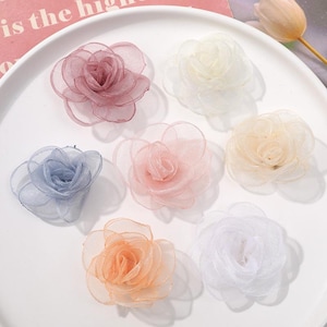 Puede incluir: Siete flores decorativas de tela dispuestas sobre un plato blanco. Las flores están hechas de material transparente y superpuesto en tonos rosa polvoriento, crema, azul claro, rosa suave, beige, melocotón y blanco, con un diseño de rosa.