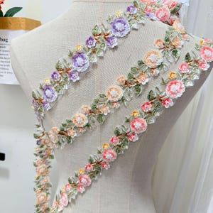 以下が含まれることがあります： 紫、ピーチ、ピンクの色合いで刺繍された花柄の装飾用トリム。緑の葉、小さな白いビーズ、透明なスパンコールがシアーなメッシュリボンに配置されており、重なり合う花のつるのようなデザインが特徴です。