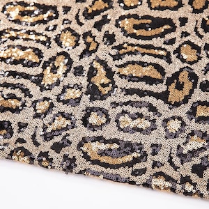 May include: A fabric surface covered in dense, shimmering sequins arranged in a leopard print pattern. The design features a repeating motif of black, gold, and silver-toned spots, creating a textured, reflective appearance across the entire material.