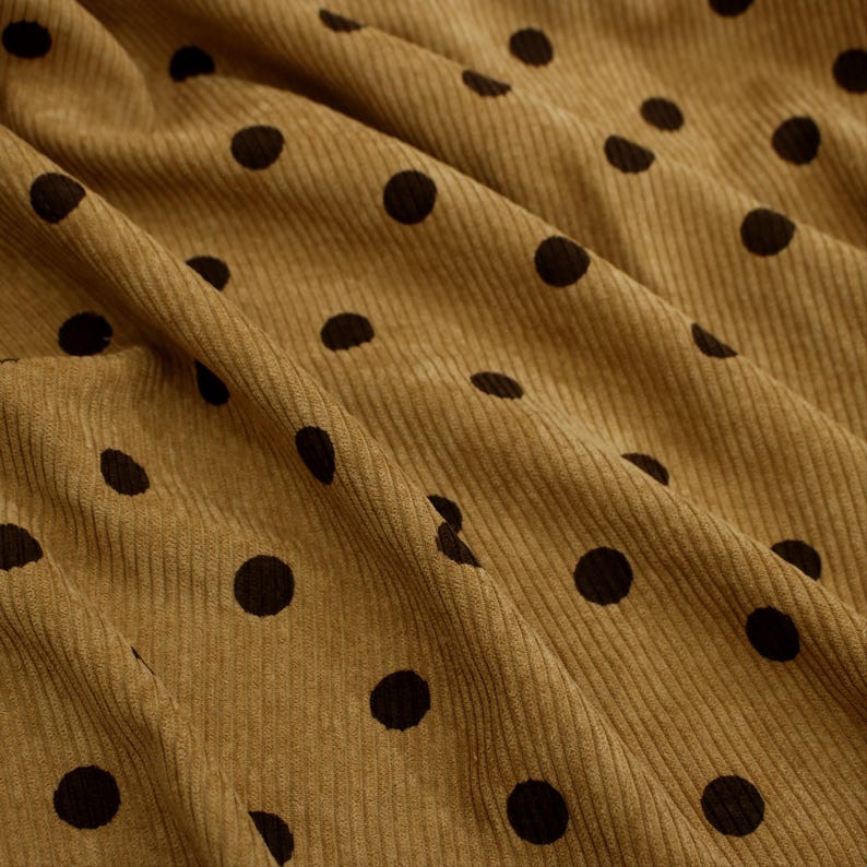 May include: A mustard yellow corduroy fabric featuring a repeating pattern of small, solid black polka dots. The material is shown with soft folds and drapes, highlighting the vertical ribbed texture characteristic of the corduroy weave.