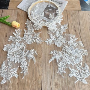 May include: A white lace applique featuring intricate floral embroidery with leaves and vines on a sheer mesh base. The design consists of two symmetrical floral clusters connected by a central circular lace element adorned with small white beads.