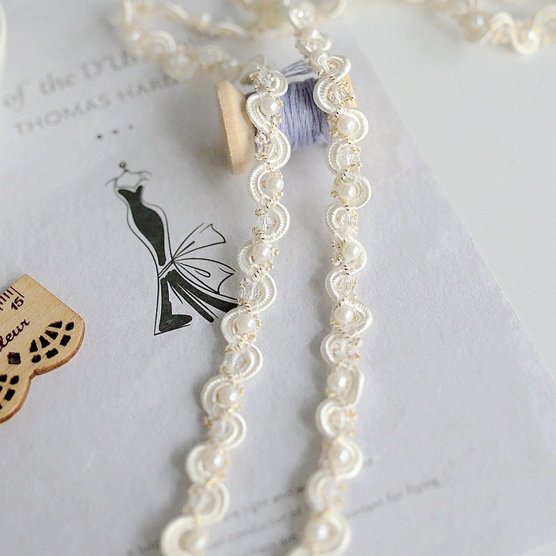 May include: White beaded trim with a scalloped edge and gold thread accents. The trim is wound around a spool.