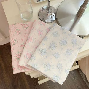 May include: Three floral fabric pieces in pink and off-white. The fabrics feature delicate rose and blue floral patterns. The textiles are folded and stacked, suggesting they are ready for use in sewing or crafting projects. The fabrics are on a white surface.