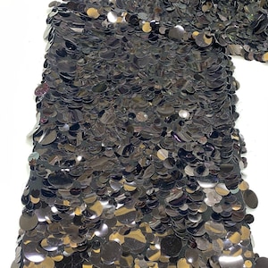 Puede incluir: Una tela negra densamente cubierta con lentejuelas circulares superpuestas. Las lentejuelas son reflectantes y oscuras, creando una superficie texturizada y brillante. La palabra Black est&aacute; impresa en la esquina superior izquierda.